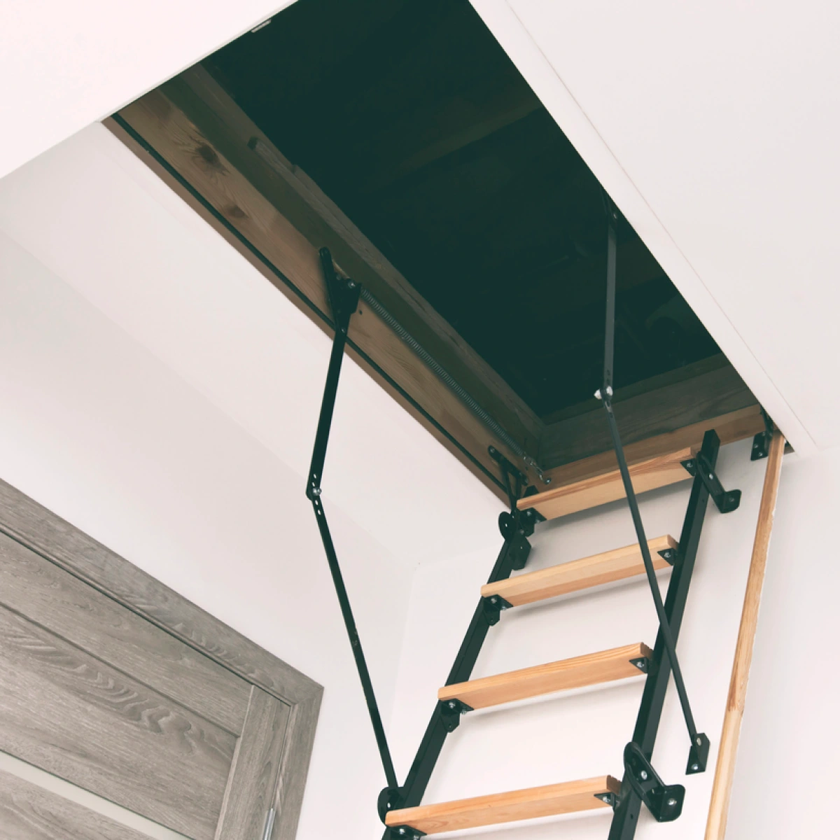 An open attic door in a white ceiling, revealing wooden steps that lead up to the attic space