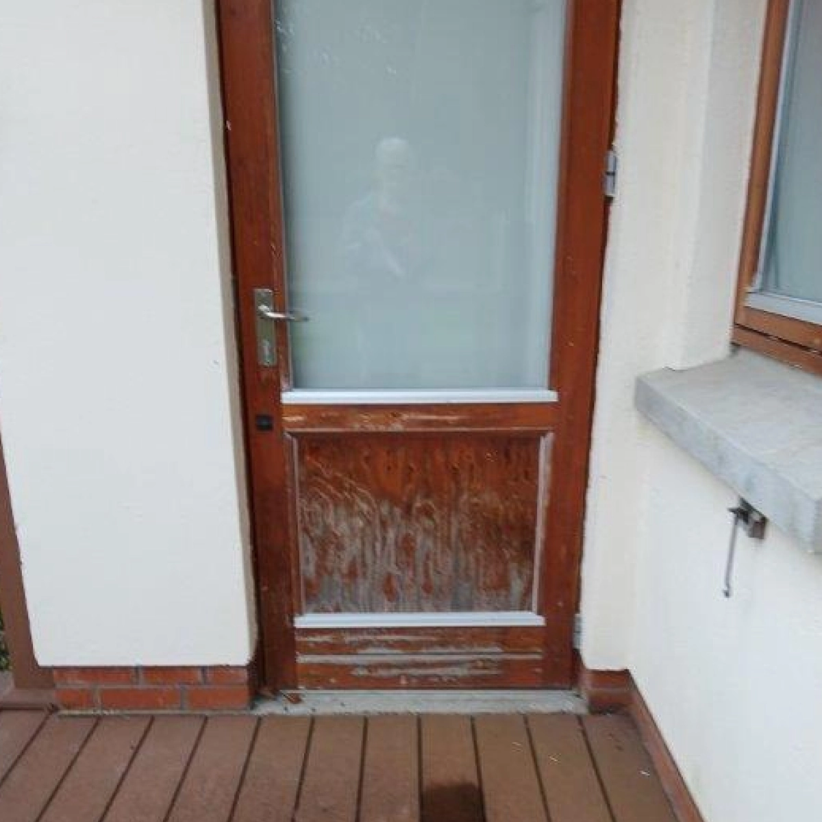 Weathered, narrow brown door with a glass pane, set in a white building. The door has an aged appearance.