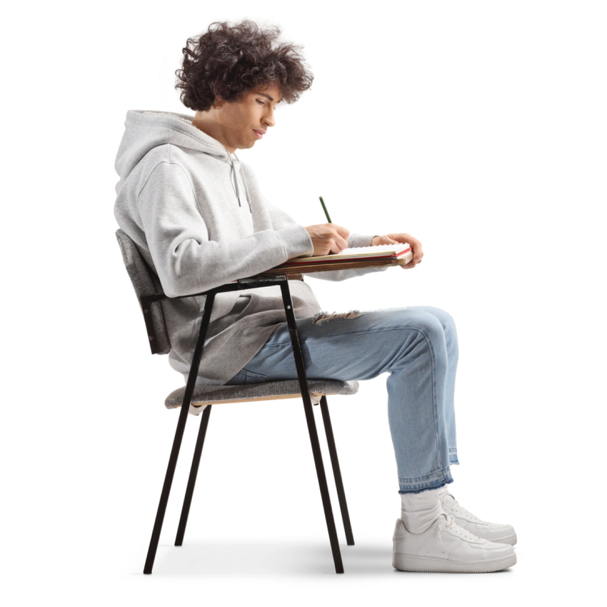 A man in a grey hoodie and blue jeans sits on an office chair with an attached table, writing in a notebook with a pen.