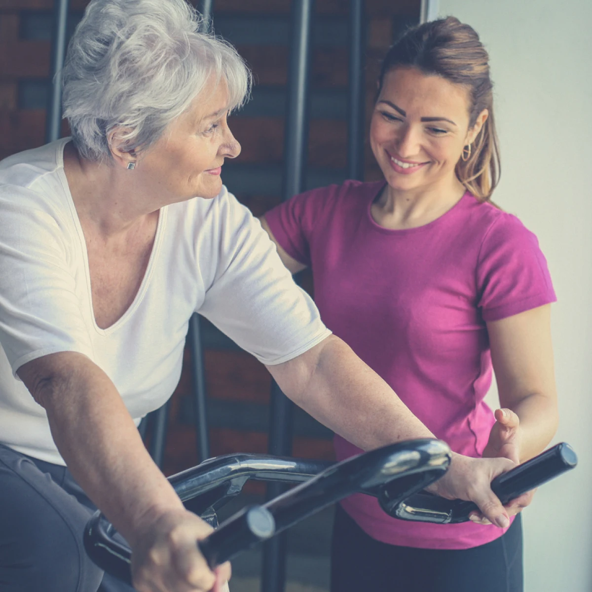 Senior woman exercising on a stationary bike in fitness class. A female trainer stands beside the bike.